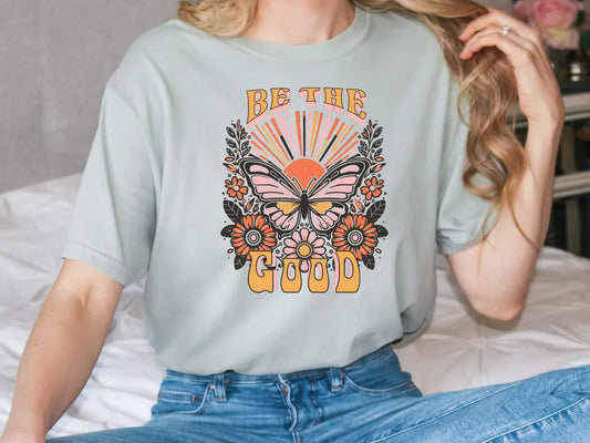 a woman sitting on a bed wearing a t - shirt that says be the good