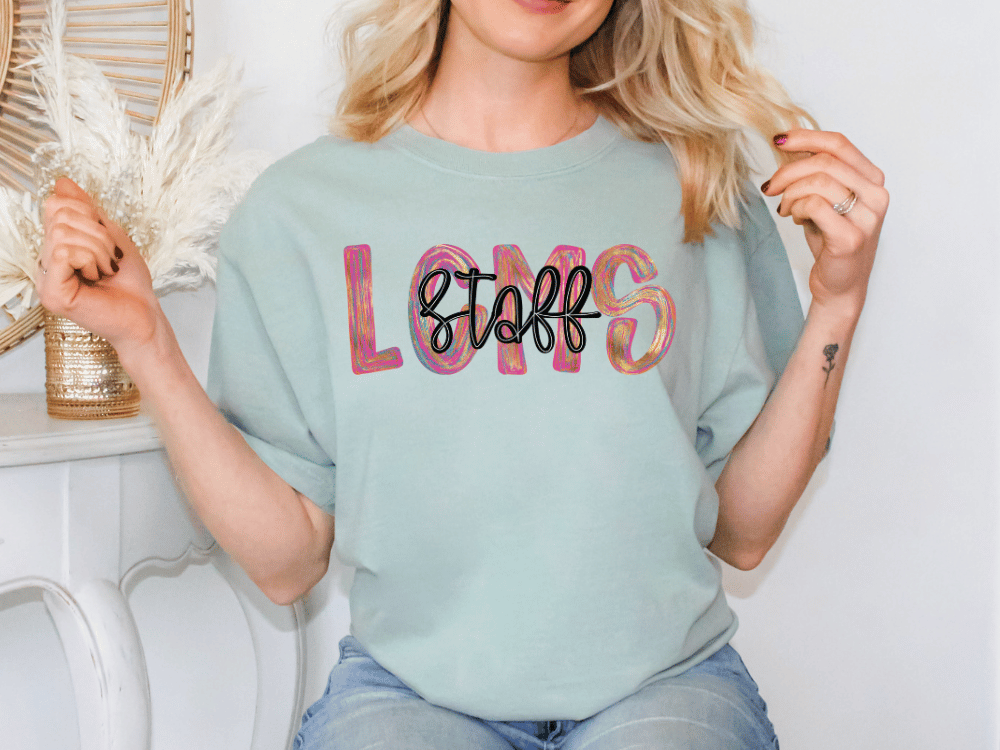 A woman wearing a light blue t-shirt with the word "LOMS" printed on it, sitting in front of a white wall.