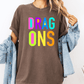 A person wearing a brown t-shirt with the text "DRAG ONS" in colorful letters, along with a denim jacket, stands against a white background.