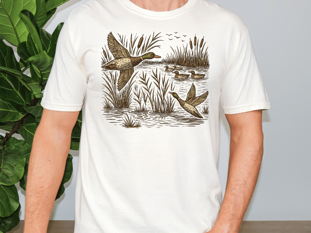 A white t-shirt with a graphic design depicting ducks swimming in a pond surrounded by reeds.