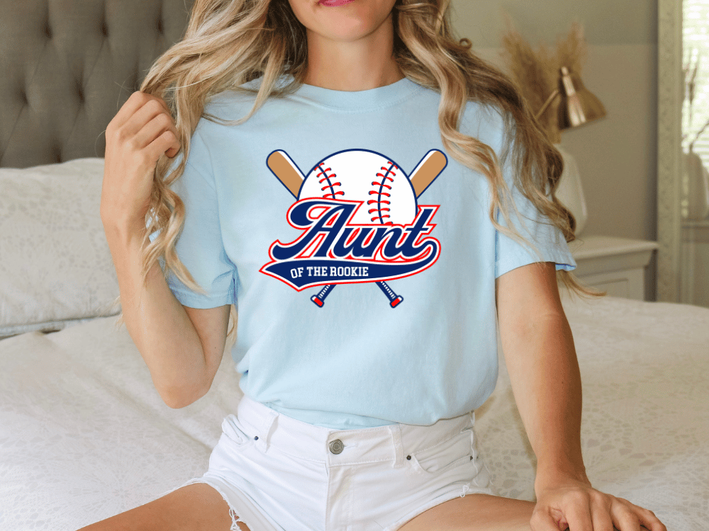 A woman wearing a light blue t-shirt with a graphic design of a baseball bat and ball, along with the text "Aunt of the Rookie".