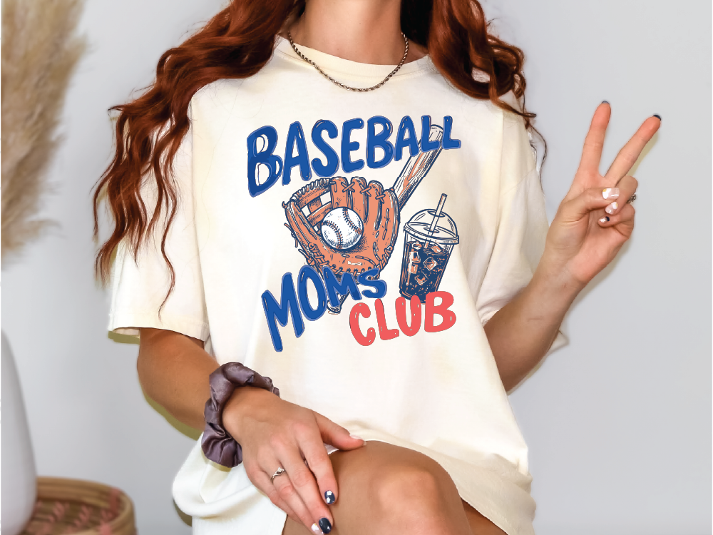 A person wearing a white t-shirt with a baseball graphic and the text "Baseball Moms Club" is making a peace sign with their hand.