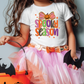 A young girl wearing a white t-shirt with a Halloween-themed design and a pink tulle skirt, holding a black bat and a pumpkin-shaped bucket.