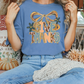 A woman wearing a blue t-shirt with a graphic design and text, sitting on a bench surrounded by fall decorations.