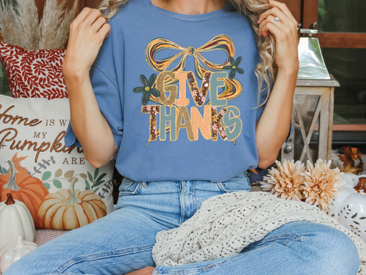 A woman wearing a blue t-shirt with a graphic design and text, sitting on a bench surrounded by fall decorations.