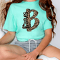 A person wearing a turquoise t-shirt with a leopard print design and gray shorts is sitting on the floor.