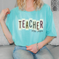 A woman wearing a teal t-shirt with the word "TEACHER" printed on it, sitting on a couch.