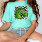 A person wearing a turquoise t-shirt with a graphic of a green dragon and leopard print accents, sitting cross-legged on the floor.
