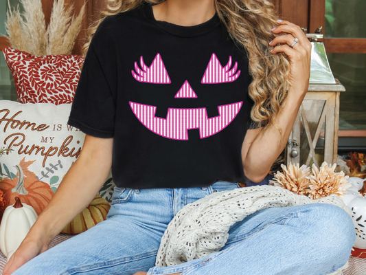 A woman wearing a black t-shirt with a pink jack-o-lantern graphic is sitting on a couch surrounded by fall decorations.