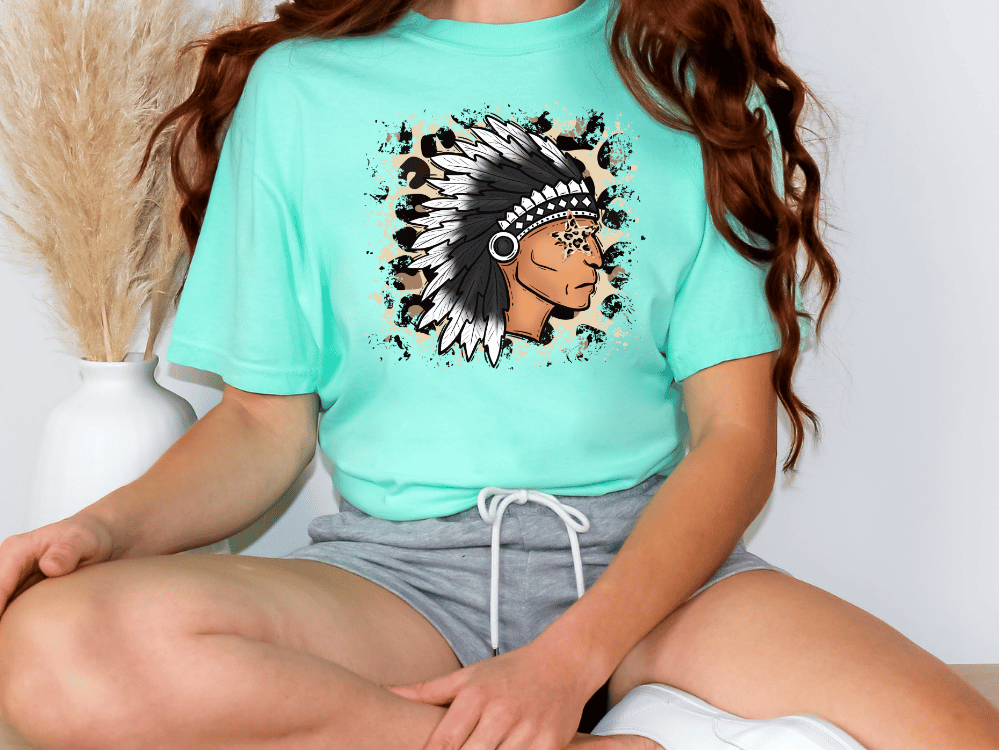 A person wearing a turquoise t-shirt with a graphic of a Native American headdress and gray shorts, sitting on the floor.