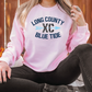 A woman wearing a pink sweatshirt with the text "LONG COUNTY BLUE TIDE" printed on it, sitting on a blanket with pumpkins in the background.