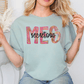 A woman wearing a light blue t-shirt with the word "MES" printed on it, sitting in front of a white wall.