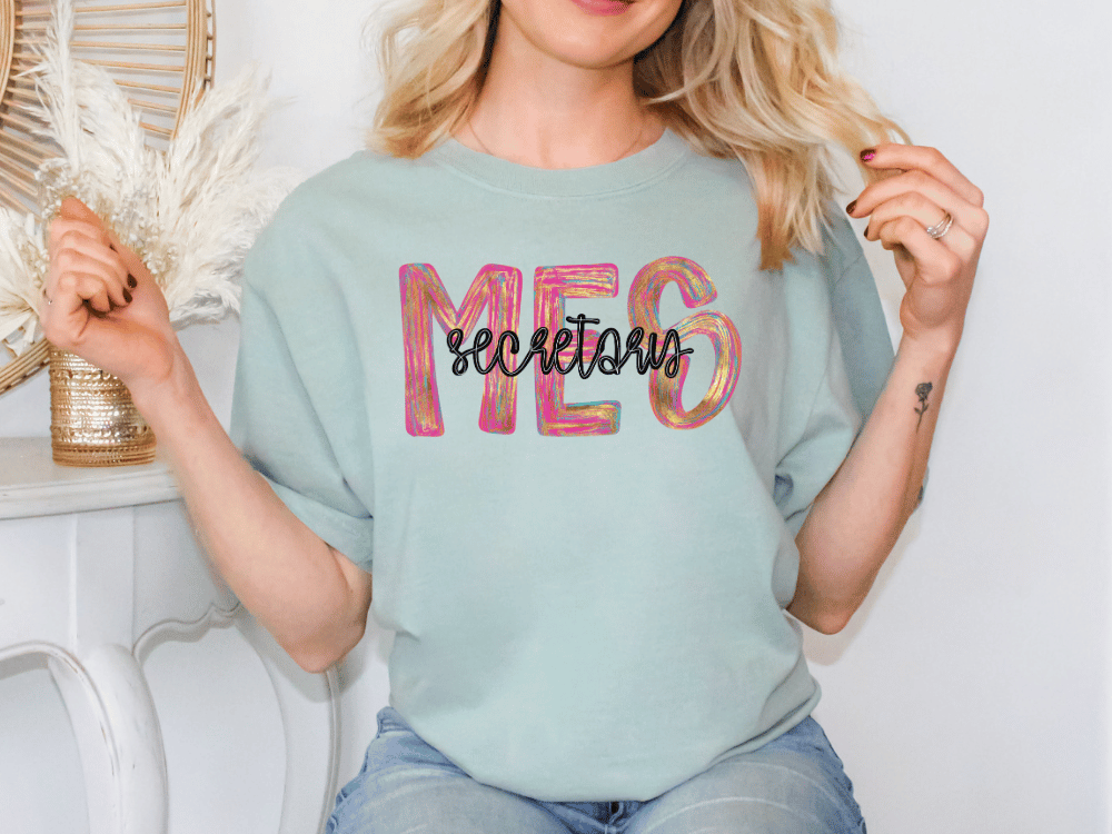 A woman wearing a light blue t-shirt with the word "MES" printed on it, sitting in front of a white wall.