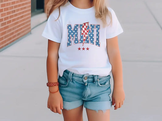 a little girl standing on a sidewalk wearing a t - shirt with an american flag