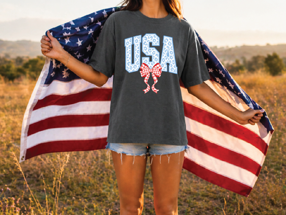 A person stands in a field holding an American flag, wearing a grey t-shirt with the word "USA" printed on it.