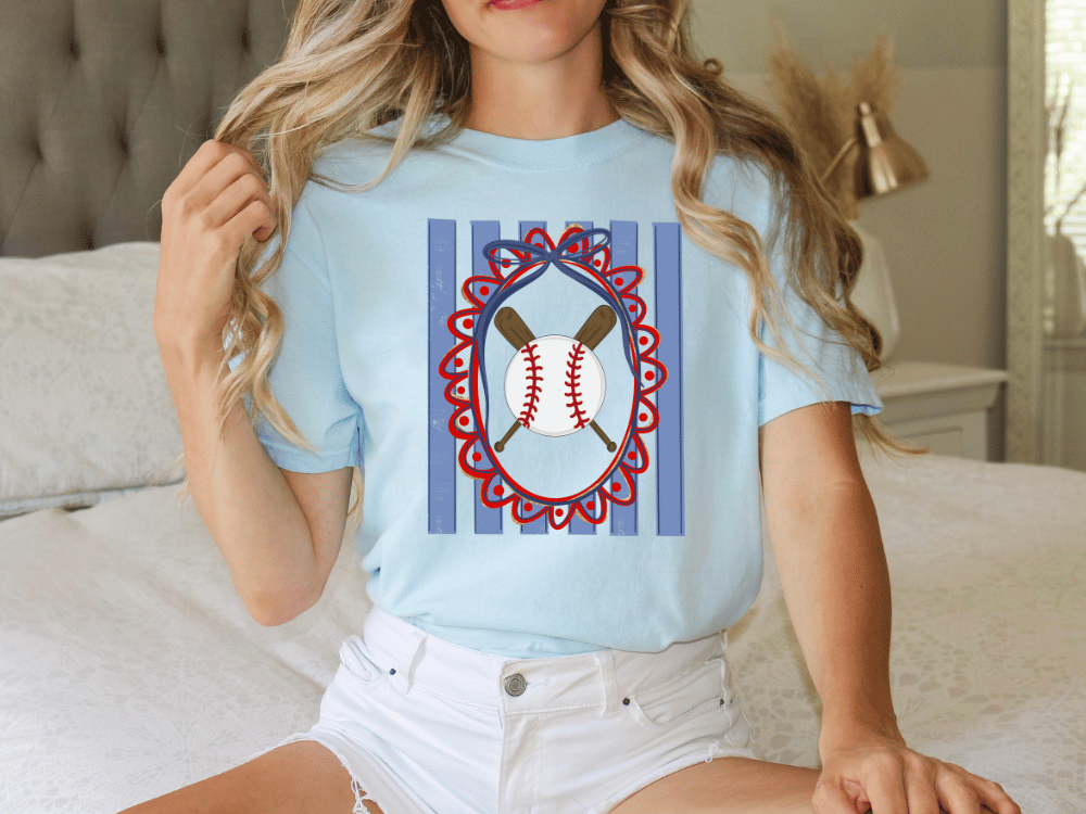 A woman wearing a light blue t-shirt with a graphic of a baseball and a heart-shaped frame is sitting on a bed.