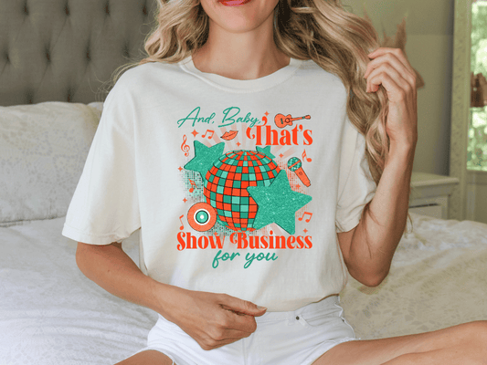 A woman wearing a white t-shirt with a graphic design featuring a disco ball, stars, and text that says "And Baby's That's Show Business for you".