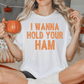 A woman sitting on a bed, wearing a white t-shirt with the text "I WANNA HOLD YOUR HAM" printed on it. She is holding a pumpkin-shaped mug and appears to be enjoying a warm beverage.