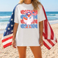 A woman wearing a white t-shirt with a graphic of a bird holding an American flag, standing on a beach with the ocean in the background.