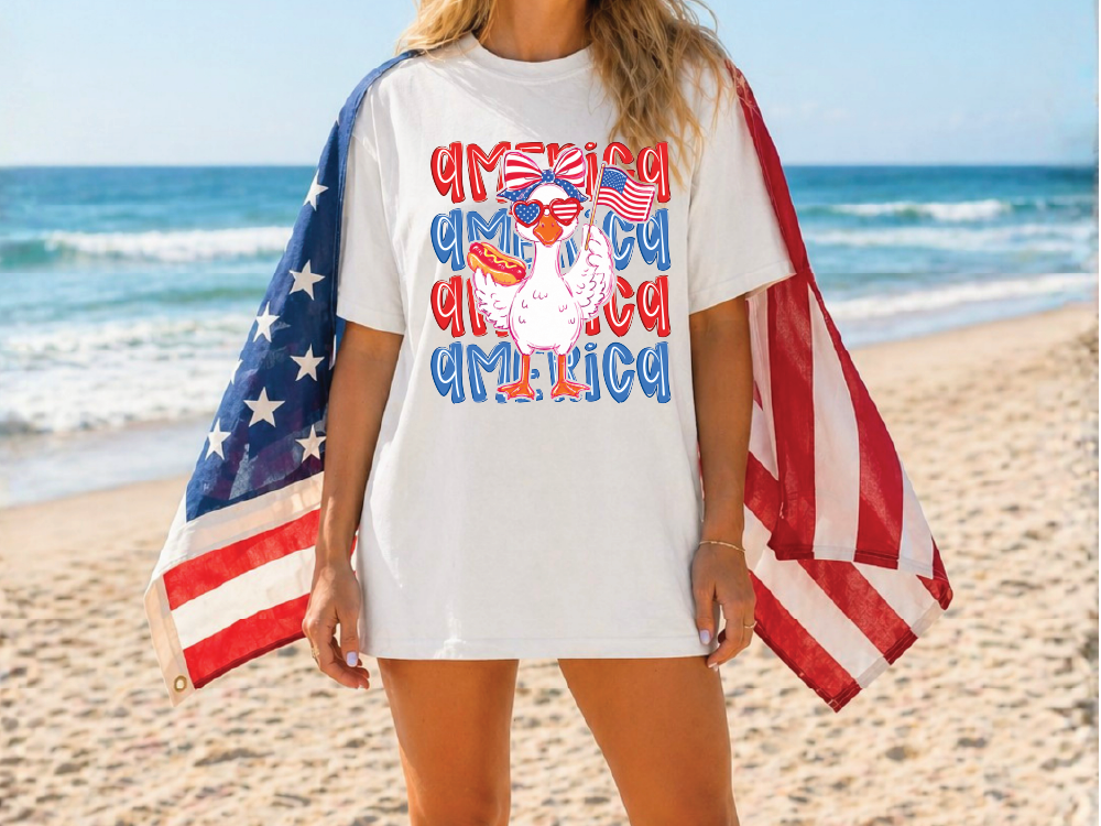 A woman wearing a white t-shirt with a graphic of a bird holding an American flag, standing on a beach with the ocean in the background.