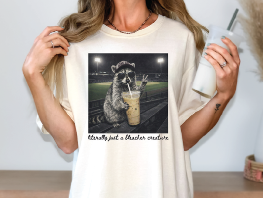 A person wearing a white t-shirt with a graphic of a raccoon holding a drink and wearing a baseball cap.