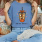 A person wearing a blue t-shirt with a graphic of a coffee drink and the text "ON THE ROCKS" is sitting on a bench surrounded by fall decorations.