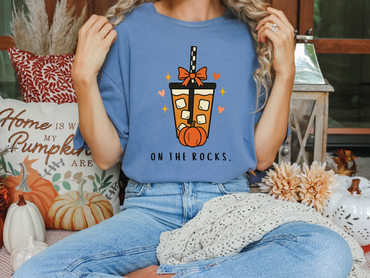 A person wearing a blue t-shirt with a graphic of a coffee drink and the text "ON THE ROCKS" is sitting on a bench surrounded by fall decorations.