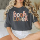 A woman wearing a black t-shirt with the text "book lover" embroidered on it, sitting on a bed.