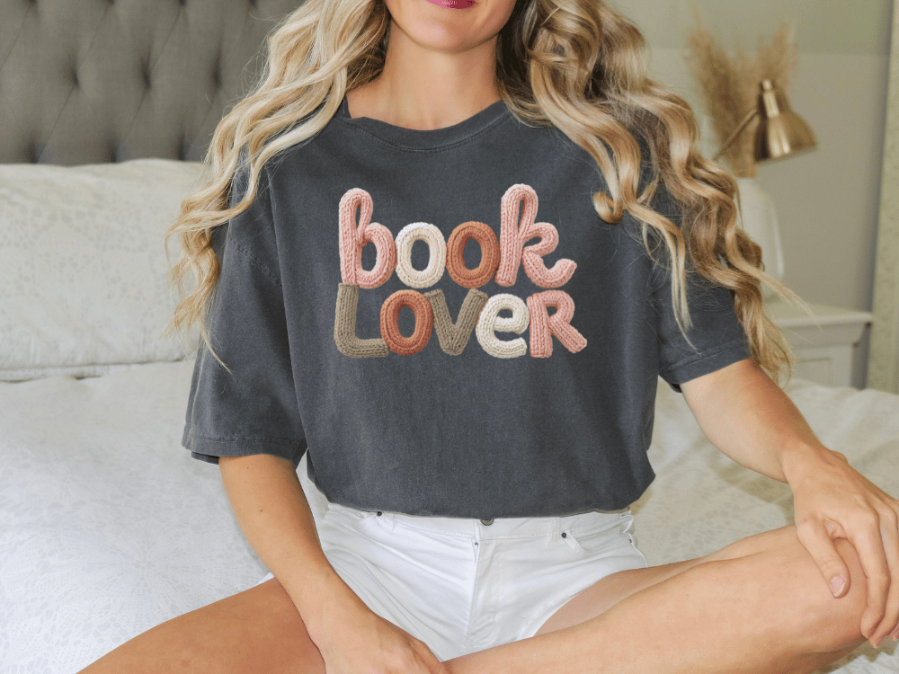 A woman wearing a black t-shirt with the text "book lover" embroidered on it, sitting on a bed.