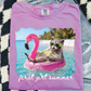 A pink t-shirt with a graphic design depicting a raccoon wearing sunglasses and holding a drink, floating on a pink flamingo-shaped inner tube in a tropical beach setting.