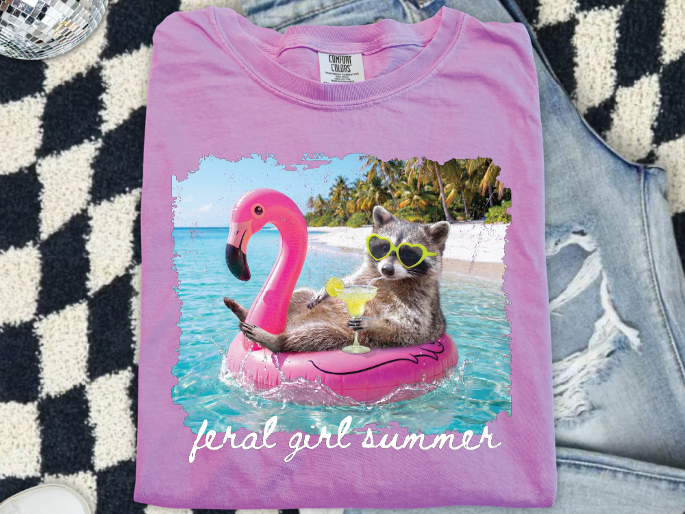 A pink t-shirt with a graphic design depicting a raccoon wearing sunglasses and holding a drink, floating on a pink flamingo-shaped inner tube in a tropical beach setting.