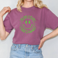 A woman wearing a purple t-shirt with a green smiley face design and blue jeans stands with her hands on her hips.