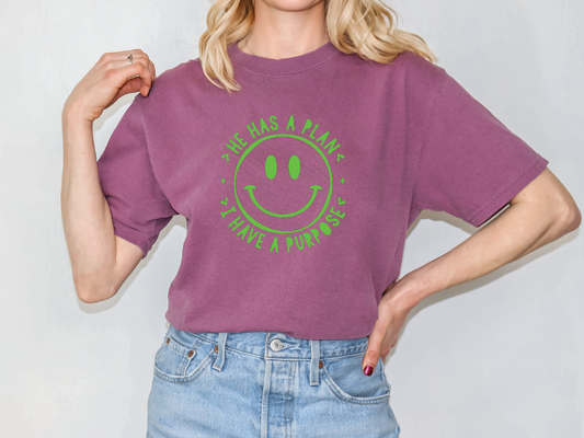 A woman wearing a purple t-shirt with a green smiley face design and blue jeans stands with her hands on her hips.