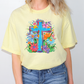A person wearing a yellow t-shirt with a colorful graphic of a cross surrounded by flowers and foliage.