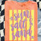 a bright orange t-shirt with the text "sun, salt & sand" printed on it, along with two purple stars. The t-shirt is placed on a checkered surface, and there is a pair of jeans visible in the background.