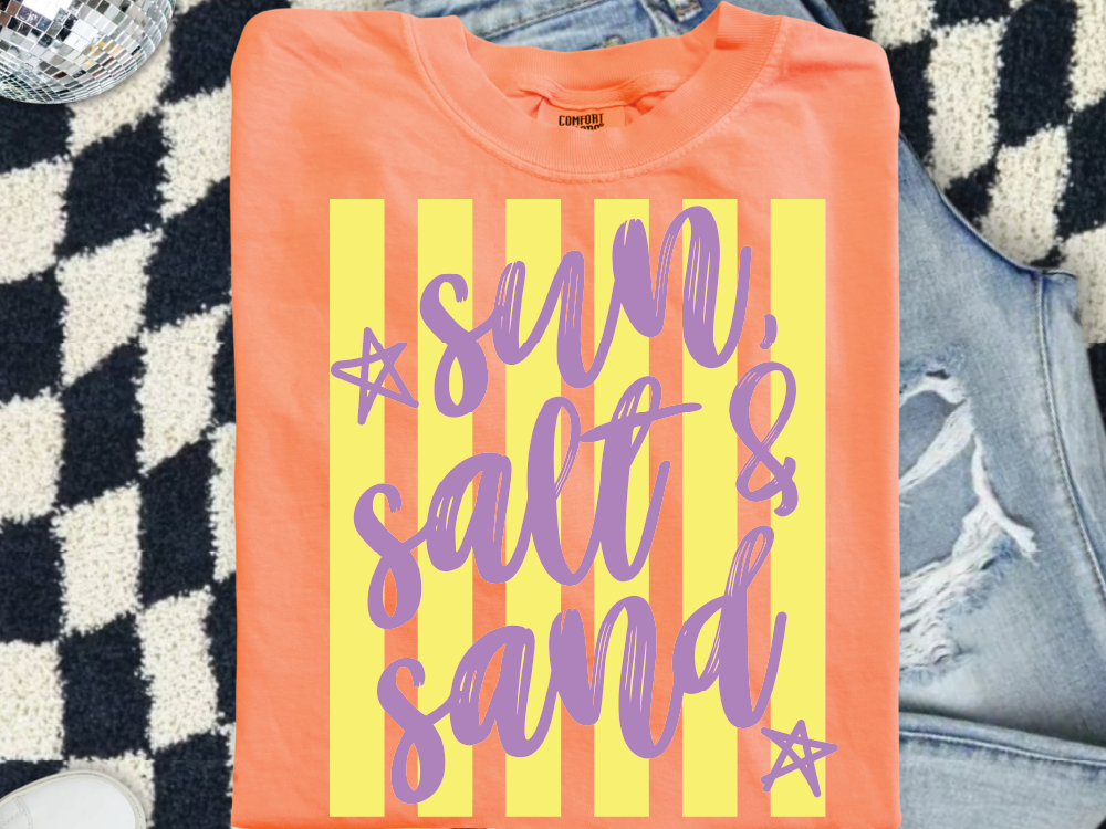 a bright orange t-shirt with the text "sun, salt & sand" printed on it, along with two purple stars. The t-shirt is placed on a checkered surface, and there is a pair of jeans visible in the background.