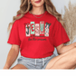 A woman wearing a red t-shirt with a Christmas-themed design and text, sitting with her legs crossed.