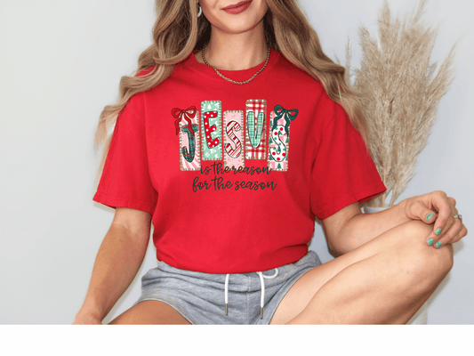 A woman wearing a red t-shirt with a Christmas-themed design and text, sitting with her legs crossed.