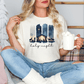 A woman sitting on a couch, wearing a white t-shirt with a nativity scene graphic and holding a mug of hot chocolate.