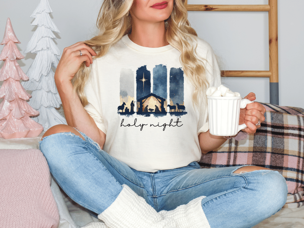 A woman sitting on a couch, wearing a white t-shirt with a nativity scene graphic and holding a mug of hot chocolate.