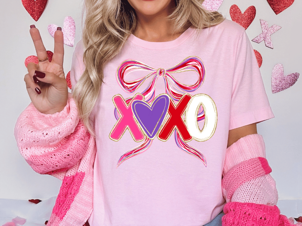 A woman wearing a pink t-shirt with a bow and hearts, making a peace sign, and pink and white striped sleeves.