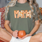A woman wearing a green t-shirt with the word "Mama" printed on it, sitting on a bed with a pumpkin-themed pillow and holding a pumpkin-shaped mug.