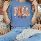A woman wearing a blue t-shirt with the text "FALL VIBES" is sitting on a bench surrounded by fall decorations, including pumpkins and a "Home is where the heart is" sign.