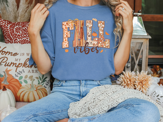 A woman wearing a blue t-shirt with the text "FALL VIBES" is sitting on a bench surrounded by fall decorations, including pumpkins and a "Home is where the heart is" sign.