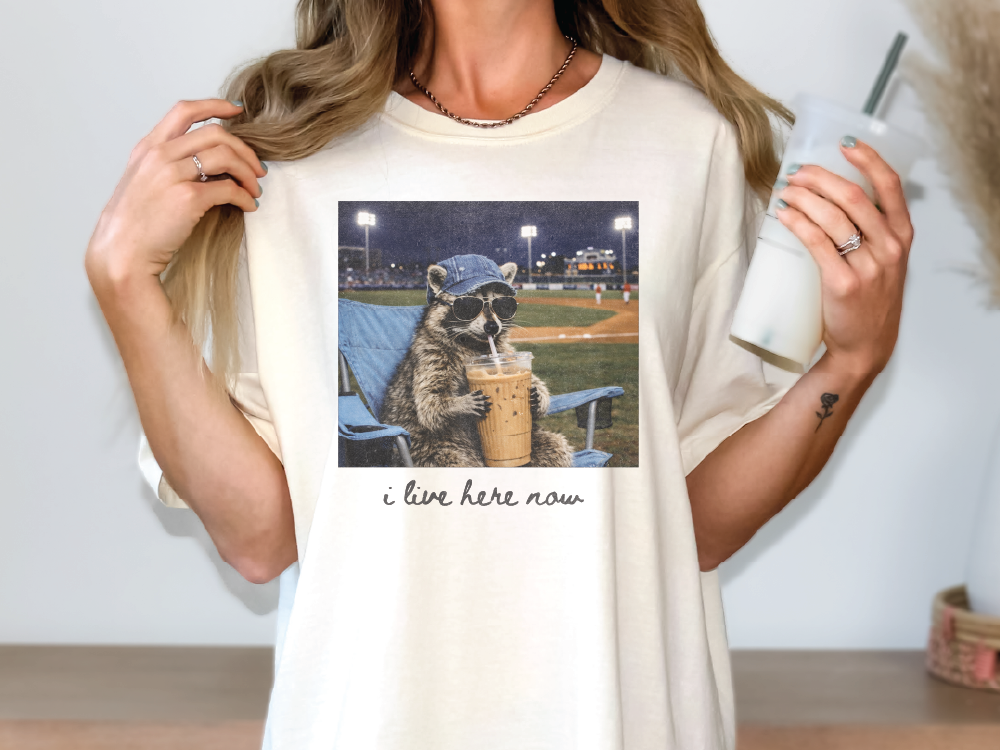 A person wearing a white t-shirt with a graphic of a raccoon sitting in a chair and drinking from a cup, with the text "I live here now" printed on the shirt.