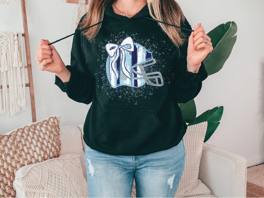 A woman wearing a black hoodie with a graphic of a football helmet and a bow on it, standing in a room with a couch and a plant in the background.