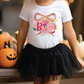 A young girl wearing a white t-shirt with a colorful graphic and a black tulle skirt stands next to a collection of carved pumpkins.