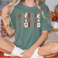 A woman wearing a green t-shirt with a graphic design sits on a bed with autumn-themed pillows and a vase of flowers.