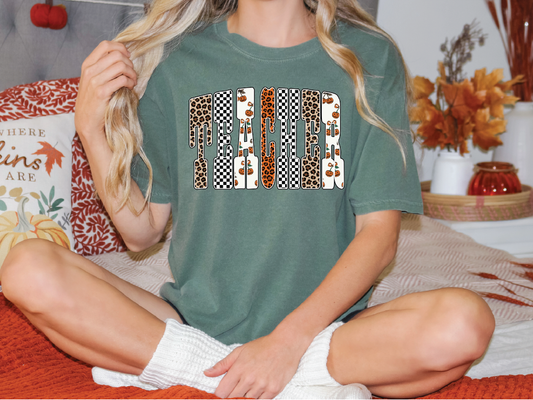A woman wearing a green t-shirt with a graphic design sits on a bed with autumn-themed pillows and a vase of flowers.
