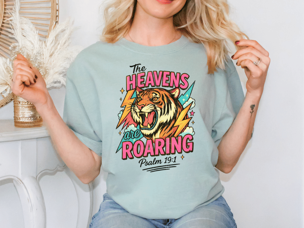 A woman wearing a light blue t-shirt with a graphic design of a tiger and the text "The Heavens are Roaring" printed on it.
